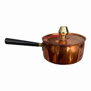 May include: A polished copper saucepan with a matching lid, featuring a long, straight black handle and a shiny gold-toned knob on the centre of the lid. The pot has a smooth, reflective metallic surface and a cylindrical shape with a flat base.