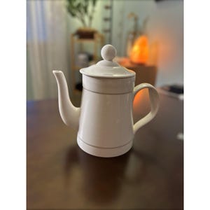 Può includere: Una teiera in ceramica bianca con coperchio e beccuccio curvo. La teiera ha un manico e un piccolo pomello sulla parte superiore del coperchio. Una sottile linea grigia circonda la teiera nella parte superiore e inferiore. La teiera è su una superficie marrone scuro.