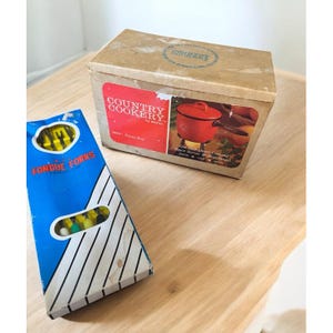 May include: A vintage Country Cookery fondue set with a red pot and a box of fondue forks. The box is tan with a red label and the words "Country Cookery" printed on it. The fondue forks box is blue with white stripes.