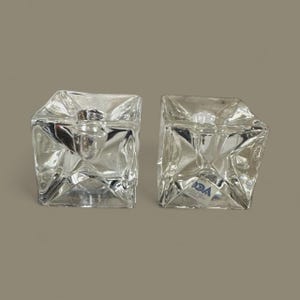 May include: Two clear glass candle holders with a geometric, faceted design. Each cube-shaped holder has a square opening at the top and a small metal insert for a candle. The holders are displayed on a neutral background.