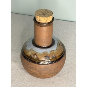 Puede incluir: Una botella de cerámica hecha a mano con un tapón de corcho. La botella tiene una base redonda y un cuello cilíndrico. La base presenta un diseño de paisaje con tonos marrones, beige y azules. El tapón de corcho es de corcho natural.