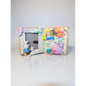 May include: A decorative Alice in Wonderland themed picture frame. The frame features a white rabbit, Alice, and the Cheshire Cat. The frame has the words "Alice in Wonderland" on it. The frame is made of a light-colored material.