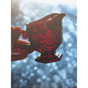 Op de afbeelding: Een robijnrode glazen creamer met een handvat en tuit. De creamer heeft een gedetailleerd, getextureerd patroon met geometrische en bloemmotieven. De creamer wordt in een hand gehouden tegen een wazige, blauw-witte achtergrond.