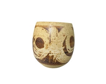 May include: A beige ceramic tumbler with a rounded shape. The tumbler features a speckled texture and is decorated with dark brown, abstract brushstroke designs. The design includes circular and triangular shapes.