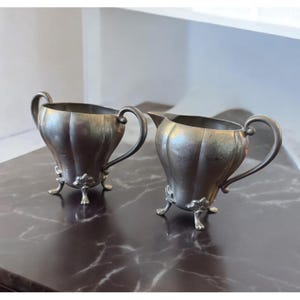 May include: Two antique silver-colored serving pieces. One is a sugar bowl with two handles, and the other is a creamer with a spout. Both have a fluted design and sit on three decorative feet. The set is displayed on a dark marble surface.