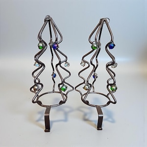 May include: Two metal tree-shaped candle holders. The holders have a dark brown finish and feature a wavy design with colorful glass beads in green, blue, and clear. The base is a circular ring with a single foot.