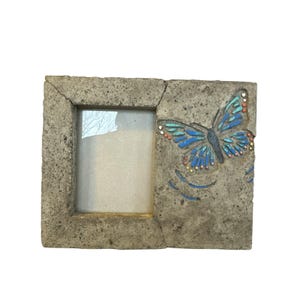 Puede incluir: Un marco de fotos rectangular con un diseño de mariposa. El marco está hecho de un material gris texturizado. Una mariposa azul con acentos coloridos está en el lado derecho. El marco tiene una abertura rectangular para una foto.