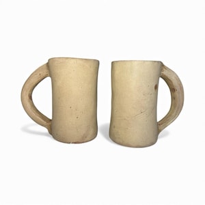 Puede incluir: Dos tazas de cerámica beige con asas curvas. Las tazas tienen forma cilíndrica y una superficie ligeramente texturizada. Las asas son gruesas y redondeadas, y las tazas están sobre un fondo blanco.