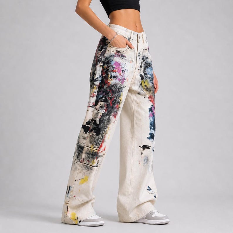 Op de afbeelding: Off-white denim broek met een wijde pijp, versierd met kleurrijke verfspatten in rood, blauw, zwart en geel. De broek heeft een klassiek ontwerp met vijf zakken en een knoop- en ritssluiting.