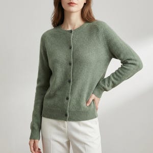 May include: A sage green cardigan with a round neckline and long sleeves. The button-down sweater is made of a soft material and has a ribbed hem and cuffs. The cardigan is paired with white pants.
