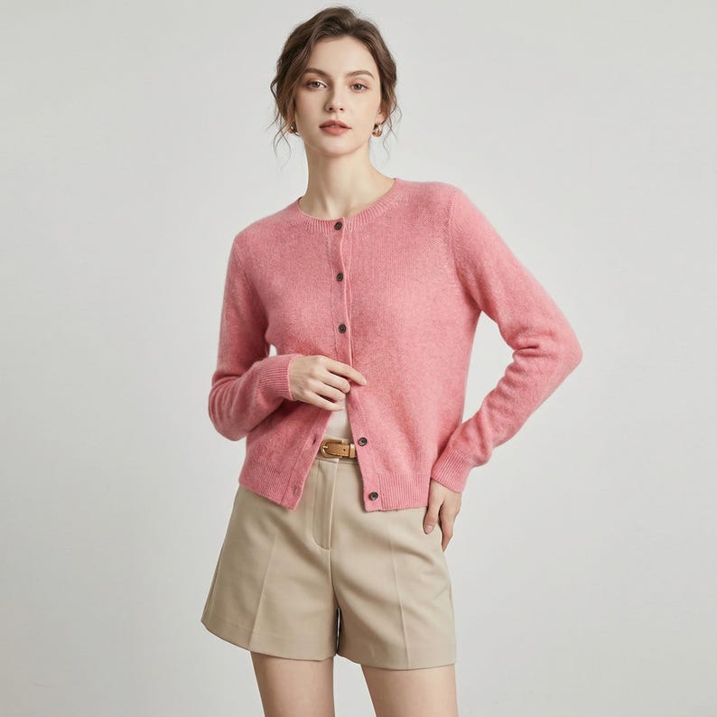 May include: A light pink cardigan with a round neckline and long sleeves. The cardigan has a button-up front and is paired with khaki shorts and a brown belt. The outfit is simple and elegant.