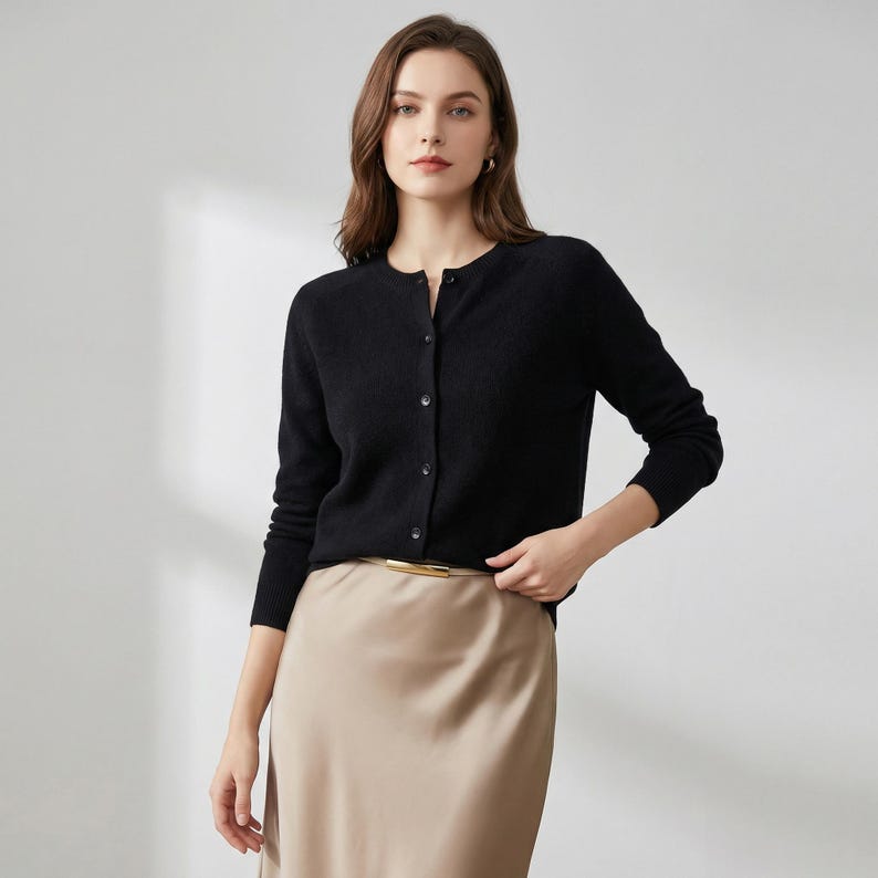 May include: A black cardigan with a round neckline and button-up front. The long-sleeved sweater is paired with a beige skirt and a gold-colored belt. The cardigan is a classic wardrobe staple.