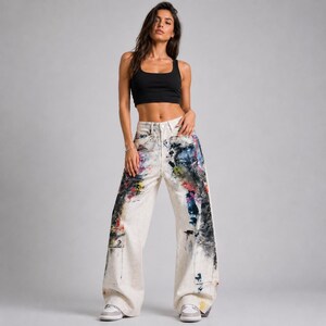 Op de afbeelding: Een persoon draagt een zwarte crop top en een witte broek met wijde pijpen. De jeans is versierd met kleurrijke verfspetters in blauw, rood, geel en zwart. De jeans heeft een hoge taille en een relaxte pasvorm.