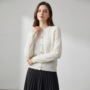 May include: A cream-colored cardigan with a round neckline and long sleeves. The sweater has a button-down front with five buttons. The person is wearing a black pleated skirt. The cardigan is a versatile piece for layering.