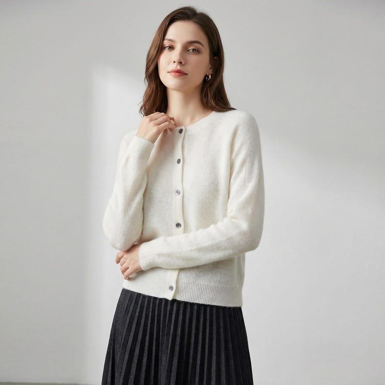 May include: A cream-colored cardigan with a round neckline and long sleeves. The sweater has a button-down front with five buttons. The person is wearing a black pleated skirt. The cardigan is a versatile piece for layering.