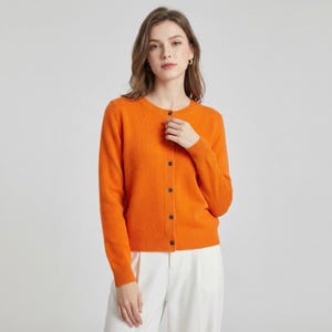 May include: An orange cardigan with a round neckline and long sleeves. The cardigan has a button-up front with dark brown buttons. The model is wearing white pants. The cardigan is made of a soft material.