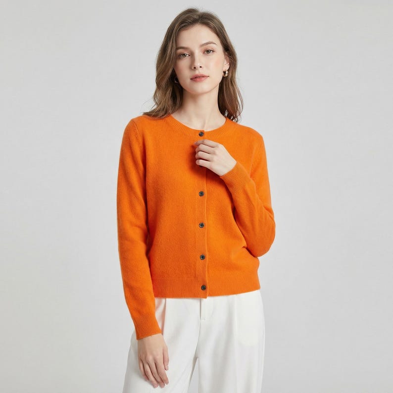 May include: An orange cardigan with a round neckline and long sleeves. The cardigan has a button-up front with dark brown buttons. The model is wearing white pants. The cardigan is made of a soft material.