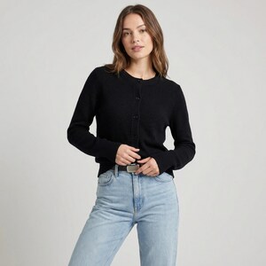 May include: A black button-down cardigan with long sleeves. The cardigan is made of a textured fabric and has a round neckline. The person is wearing light blue jeans and a silver belt.