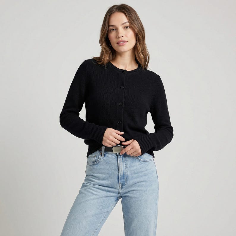 May include: A black button-down cardigan with long sleeves. The cardigan is made of a textured fabric and has a round neckline. The person is wearing light blue jeans and a silver belt.