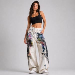 Op de afbeelding: Een vrouw draagt een zwarte tanktop en een off-white broek met wijde pijpen. De jeans is versierd met kleurrijke verfspatten in roze, blauw, geel en zwart. De jeans heeft een hoge taille en een relaxte pasvorm.