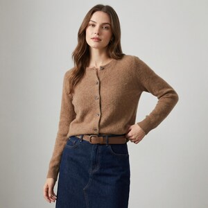 May include: A light brown cardigan with a round neckline and button closure. The cardigan is paired with a brown belt and a dark blue denim skirt. The outfit is simple and classic.
