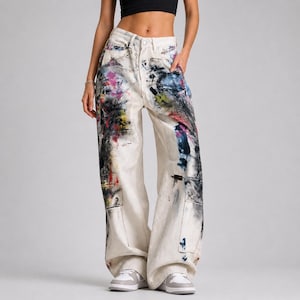 Op de afbeelding: Off-white denim jeans met een wijde pijp en een hoge taille. De jeans zijn bespat met kleurrijke verf in tinten roze, blauw, zwart en rood. Kenmerken zijn twee voorzakken en twee cargozakken. Het model draagt witte sneakers.