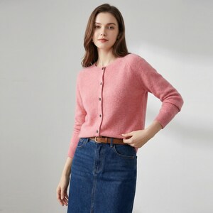 May include: A light pink cardigan with long sleeves and a round neckline. The cardigan has a button-up front and is paired with a denim skirt and a brown belt. The outfit is casual and suitable for everyday wear.
