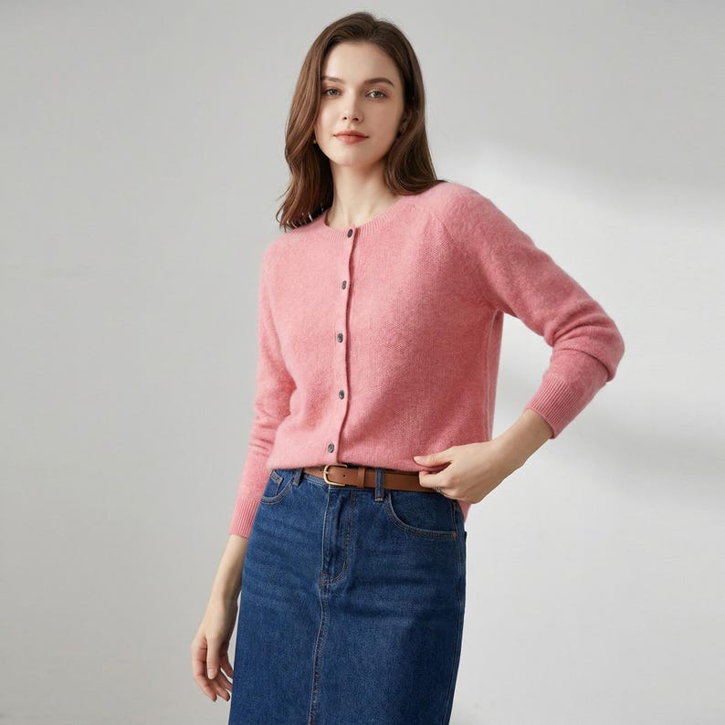 May include: A light pink cardigan with long sleeves and a round neckline. The cardigan has a button-up front and is paired with a denim skirt and a brown belt. The outfit is casual and suitable for everyday wear.