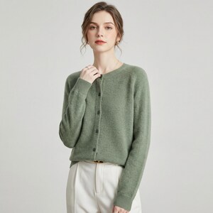 May include: A sage green cardigan with a round neckline and long sleeves. The button-down sweater is paired with white pants. The cardigan is made of a soft material and is a versatile piece for any wardrobe.