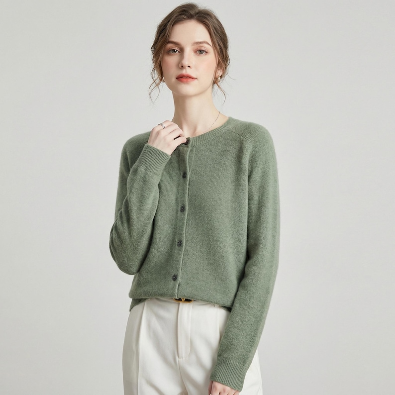 May include: A sage green cardigan with a round neckline and long sleeves. The button-down sweater is paired with white pants. The cardigan is made of a soft material and is a versatile piece for any wardrobe.