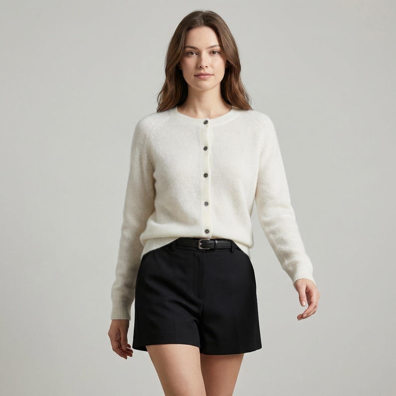 May include: A woman wearing a cream-colored cardigan with black buttons and black shorts. The shorts are held up by a black belt. The outfit is simple and classic.