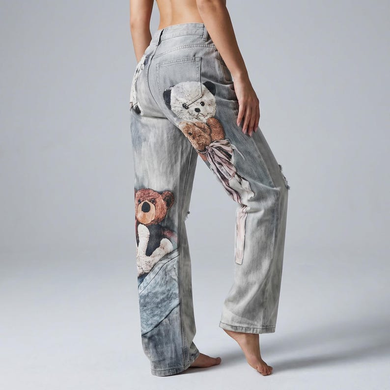 May include: Light wash denim jeans featuring a distressed design and teddy bear graphics. The jeans have a relaxed fit and are adorned with multiple teddy bear illustrations in shades of brown, white, and black. The jeans have a classic five-pocket design.