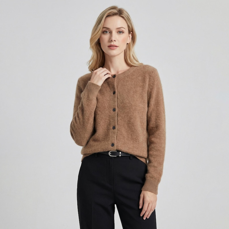 May include: A button-down, light brown cardigan sweater. The sweater has long sleeves and a round neckline. The model is wearing black pants and a black belt with a silver buckle. The sweater is made of a soft, fuzzy material.