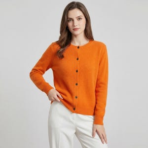 May include: An orange cardigan with long sleeves and a round neckline. The cardigan has a button-up front with dark buttons. The model is wearing white pants. The cardigan is a solid color and appears to be made of a soft material.