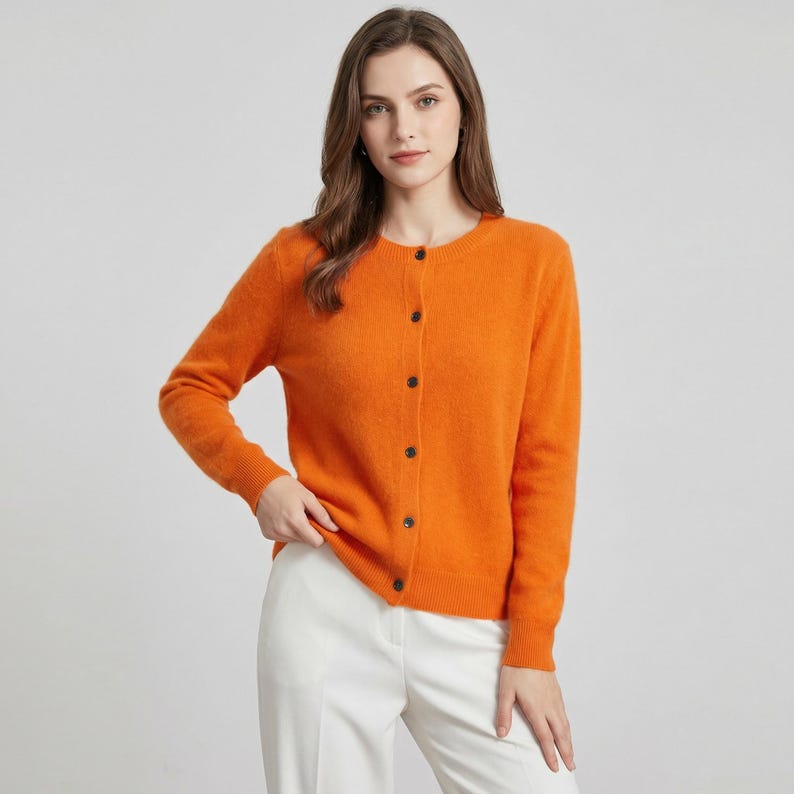 May include: An orange cardigan with long sleeves and a round neckline. The cardigan has a button-up front with dark buttons. The model is wearing white pants. The cardigan is a solid color and appears to be made of a soft material.