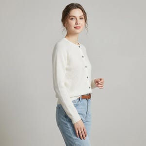 May include: A white, long-sleeved cardigan with a round neckline and button closure. The cardigan is paired with light blue jeans and a brown belt. The outfit is simple and classic.