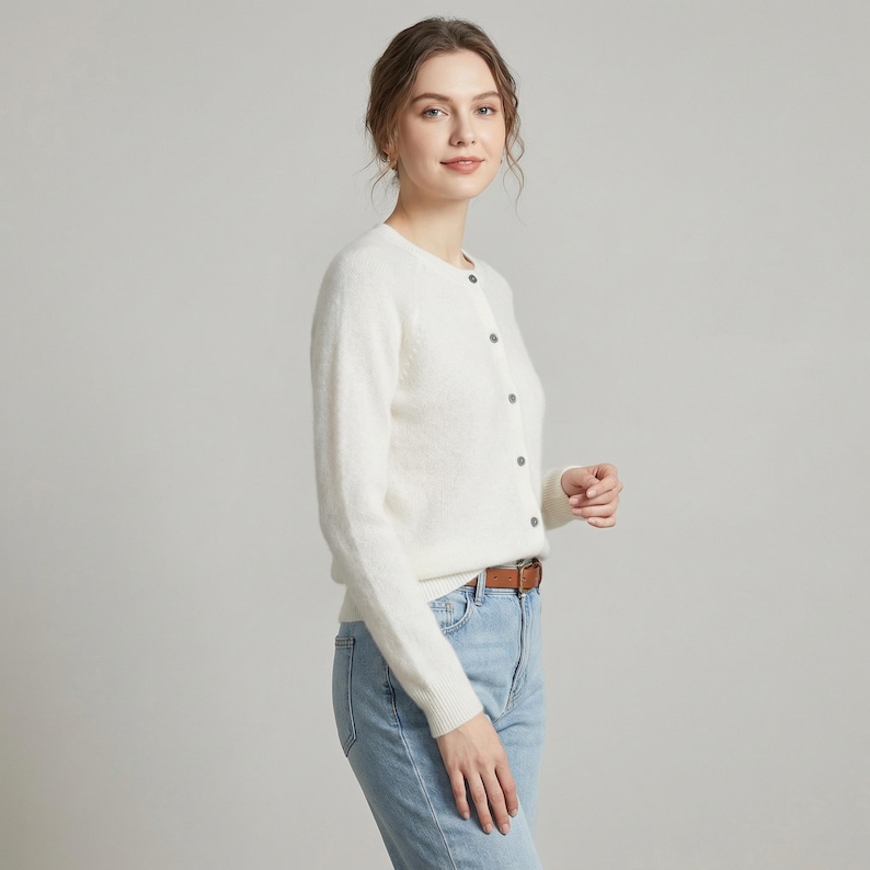 May include: A white, long-sleeved cardigan with a round neckline and button closure. The cardigan is paired with light blue jeans and a brown belt. The outfit is simple and classic.