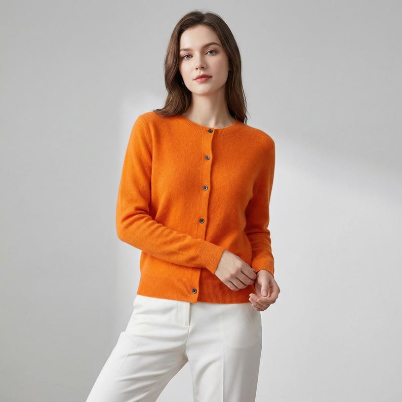 May include: An orange cardigan with a round neckline and long sleeves. The cardigan has a button-up front and is paired with white pants. The clothing is made of a soft material.