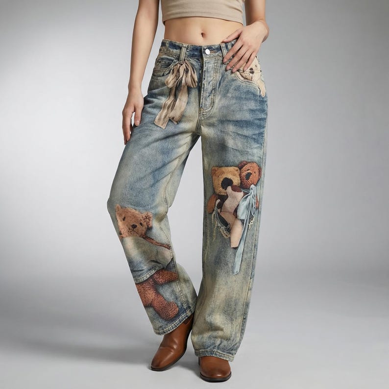 May include: Light blue denim jeans with a faded wash and a relaxed fit. The jeans feature multiple teddy bear appliqu&eacute;s in shades of brown and tan. A beige ribbon is tied at the waist. The jeans are worn with brown leather shoes.