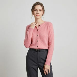 May include: A light pink cardigan with a round neckline and button-up closure. The long-sleeved sweater is paired with dark gray trousers and a black belt. The outfit is simple and elegant.