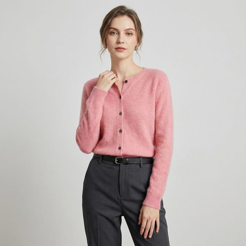 May include: A light pink cardigan with a round neckline and button-up closure. The long-sleeved sweater is paired with dark gray trousers and a black belt. The outfit is simple and elegant.