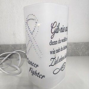 Könnte beinhalten: Weißer, mattierter Glas-Kerzenhalter mit einem Schleifendesign aus funkelnden Strasssteinen. Auf der Vorderseite steht "Cancer Fighter". Der Kerzenhalter hat Text auf Deutsch.