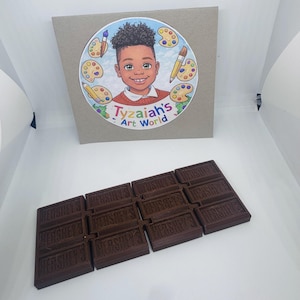 May include: A collection of dark brown Hershey's chocolate bars arranged in a rectangular shape. Above the chocolate is a round sticker with a cartoon illustration of a smiling child surrounded by art supplies and the text "Tyzai's Art World."