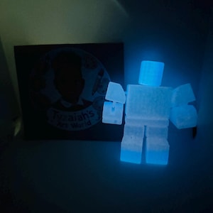 May include: A glowing blue, block-shaped robot figure with a rectangular head, arms, and legs. The robot is illuminated, giving off a bright blue glow. Beside the robot is a dark-colored square with the text "Tyzaiha's Art World".