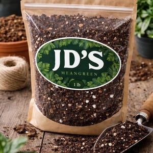 May include: A brown paper bag with a clear plastic window filled with soil. The bag has a green and white oval label that reads "JD's Meangreen" and "1 lb". A small metal trowel is filled with soil, and plants are in the background.