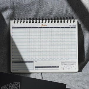 May include: A spiral-bound baseball scorebook with a white cover and grid-lined pages. The top of the book has the word "Bottom" in a cursive font. The book is open, displaying the scoring sections for a baseball game.