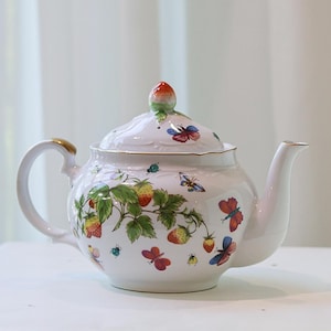 May include: A white porcelain teapot featuring a colorful pattern of red strawberries, green leaves, and various butterflies in shades of blue, red, and yellow. The lid is topped with a small strawberry-shaped finial and has gold trim.