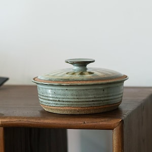 May include: A round ceramic lidded bowl featuring a speckled light blue and green glaze with a subtle sunburst pattern on the lid. The base of the bowl and the lid rim reveal the natural brown clay texture, and the lid has a small knob handle.