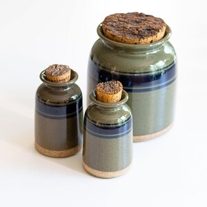 May include: A set of three ceramic storage jars with cork stoppers. The jars feature a glossy olive green glaze with a dark blue horizontal band around the upper section and a natural, unglazed tan base. The jars vary in size, with one large jar.