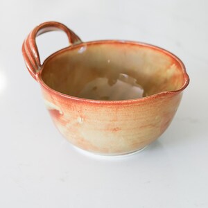 May include: A ceramic bowl with a single handle and a small pouring spout. The bowl features a warm, mottled glaze with blended tones of orange, rust, and cream, creating a smooth, glossy finish across the interior and exterior surfaces.